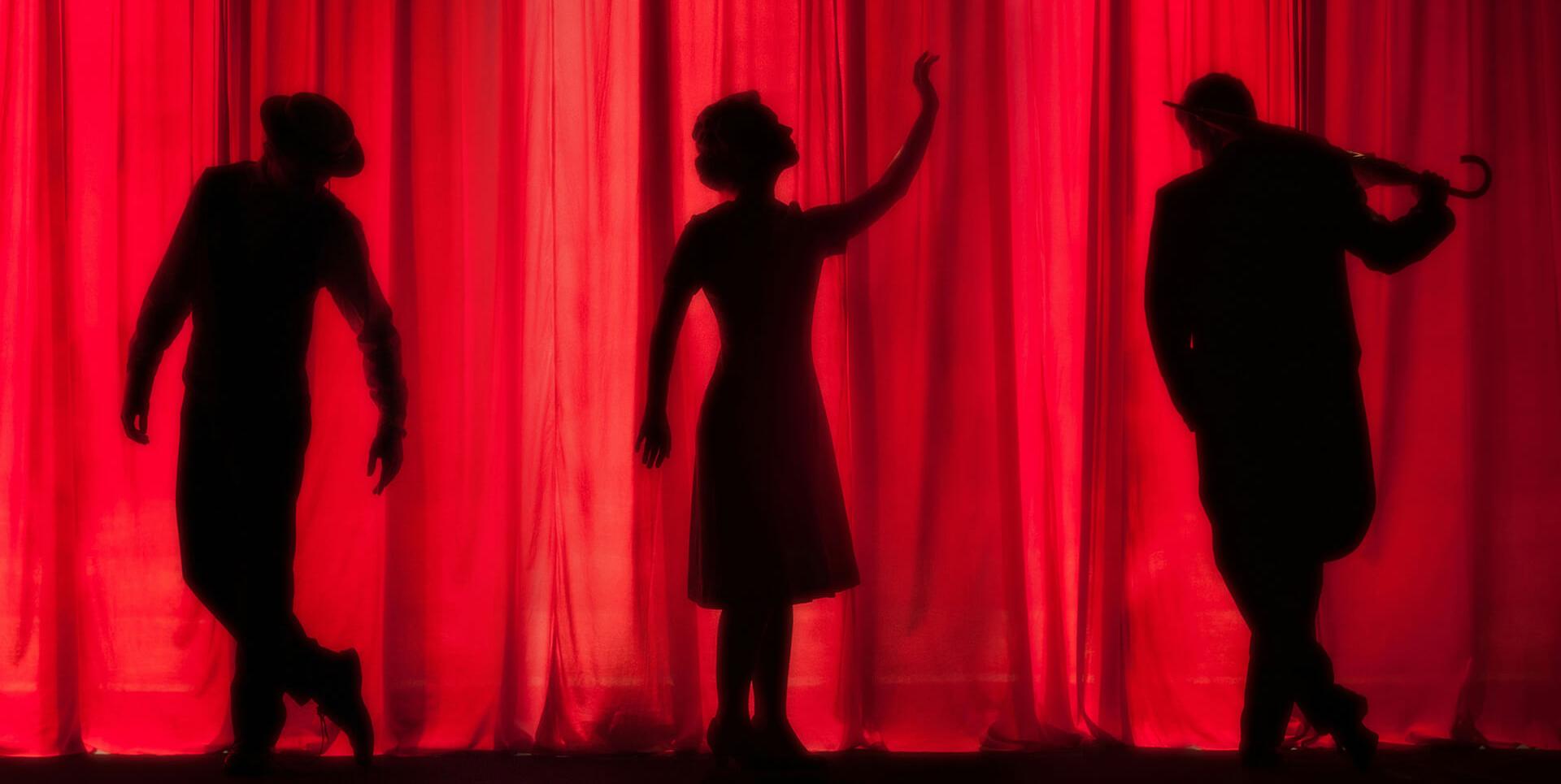 Drei dunkle Silhouetten von Darstellern auf einer Bühne vor einem roten Theatervorhang